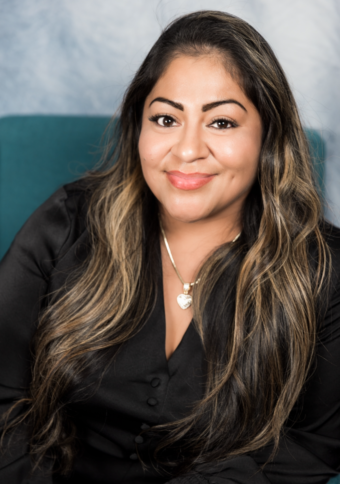 Woman with long wavy brown hair, wearing a black shirt, smiling, in front of a teal backdrop.