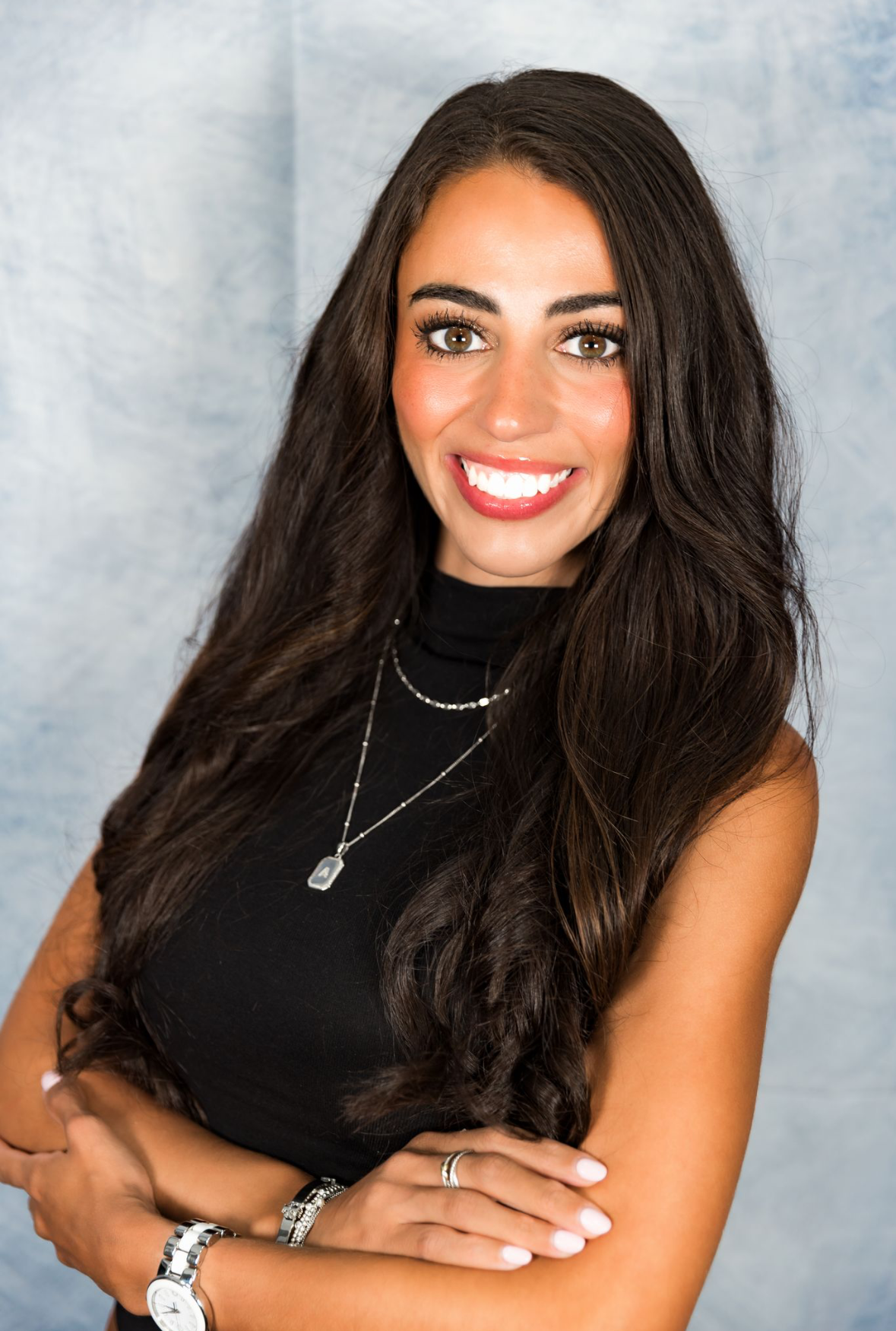 Woman with long dark hair, arms crossed, smiles at camera. Wearing a black top and jewelry.