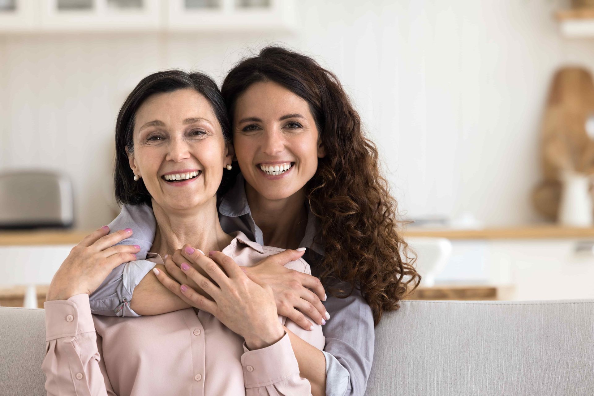 Woman hugs older woman, both smiling on a sofa indoors.