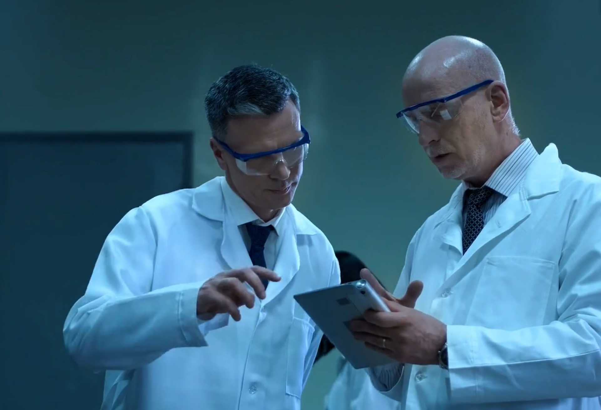 Two men in lab coats and goggles are looking at a tablet.