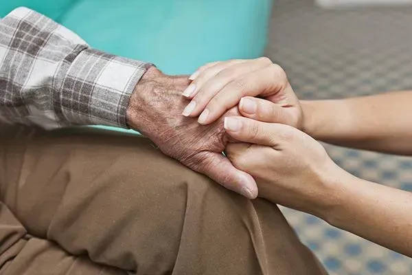 Two hands clasped in comfort, one elderly hand resting on a knee