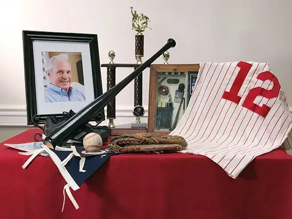 Baseball memorial display with framed photo, bat, ball, trophies, and a white jersey with red 12 on a red tablecloth