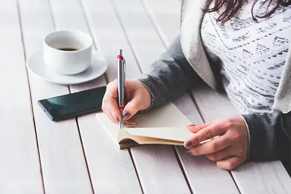 Person writing in a notebook at a white table with a coffee cup and smartphone nearby