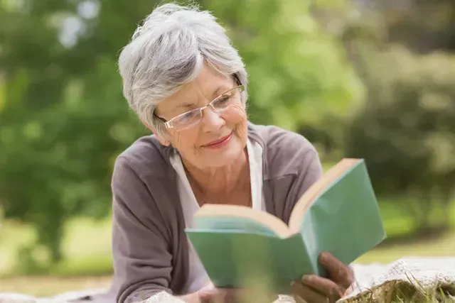 Older woman reading a book outdoors in a green garden, wearing glasses and a gray sweater
