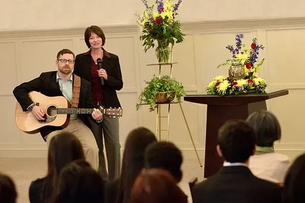 Musicians performing indoors, with a man playing guitar beside a singer and floral arrangements onstage