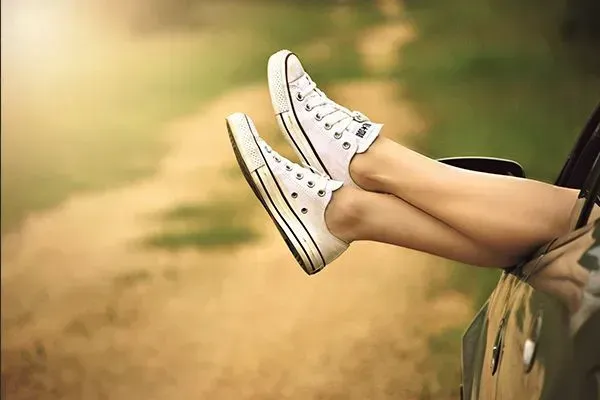 White sneakers with crossed legs sticking out of a car window on a sunny road.