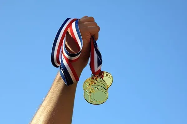 Hand holding gold medals with red, white, and blue ribbons against a blue sky