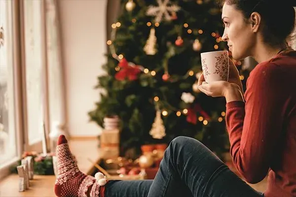 Person sitting by a Christmas tree, holding a mug and looking out a window.