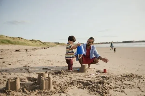 Child and adult building a sandcastle on a sunny beach, with the ocean in the background
