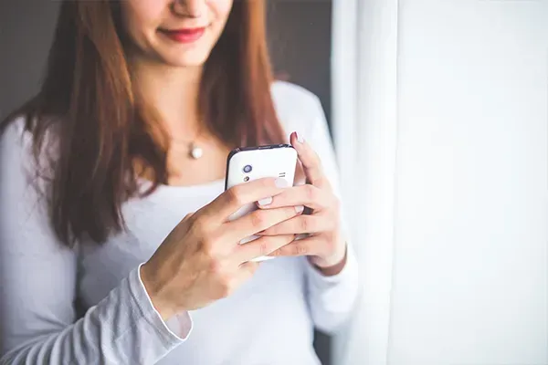 Woman holding a smartphone near a window, wearing a white top.