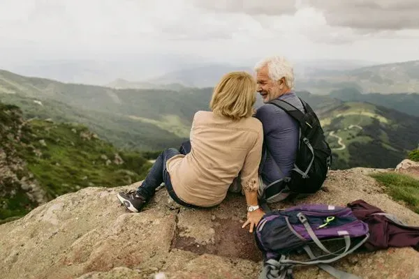 Two hikers sit on a rocky overlook, gazing at a misty mountain valley with backpacks beside them.