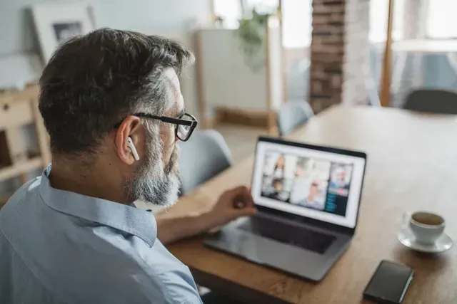 Man viewing a laptop video call at a wooden table with a coffee cup nearby
