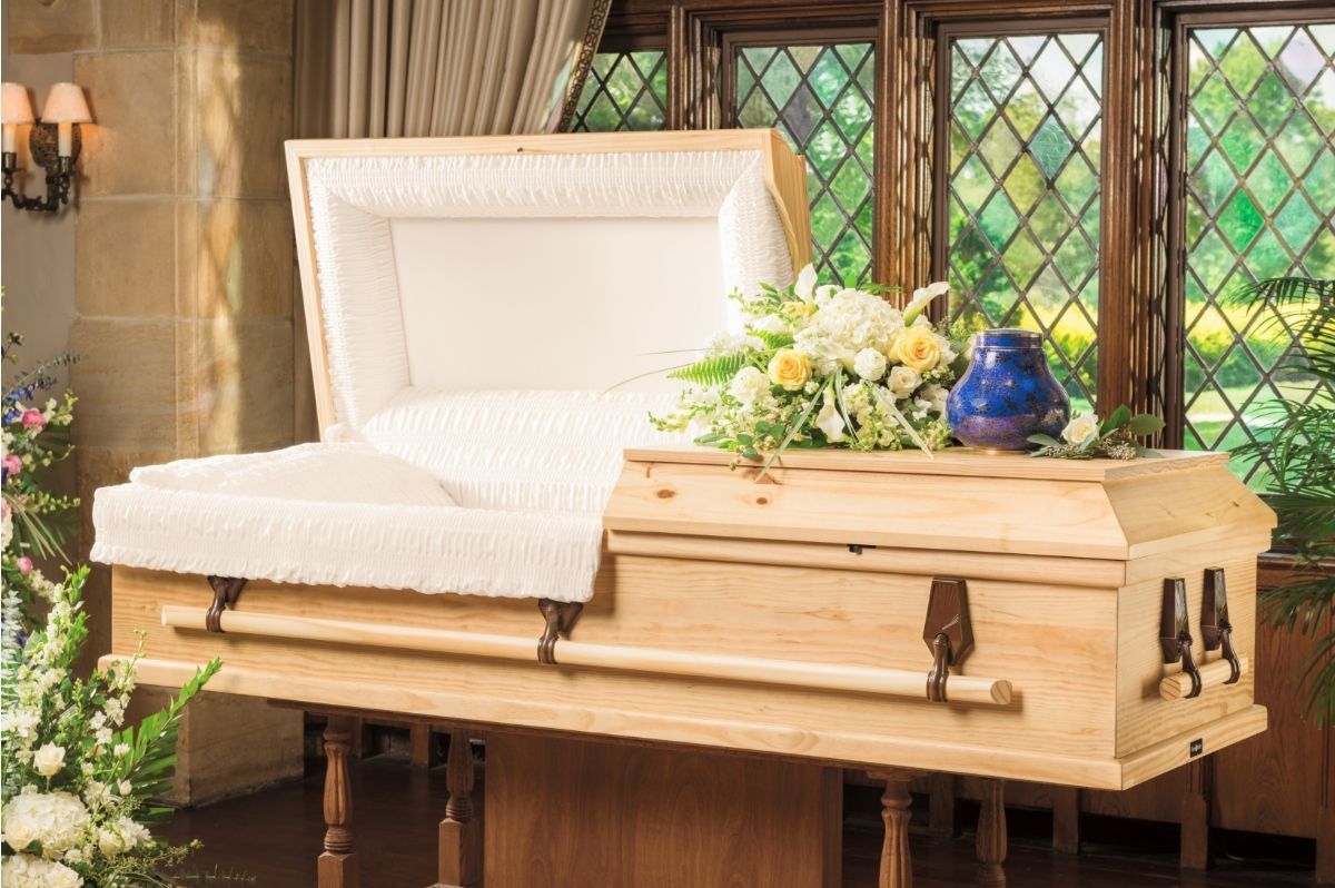 White cushioned casket with flowers in a chapel setting, viewed beside stained glass windows