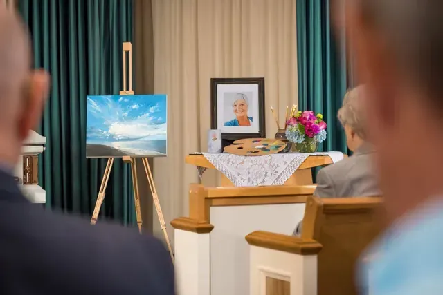 Memorial service with portrait, flowers, and a painting on an easel beside a casket.