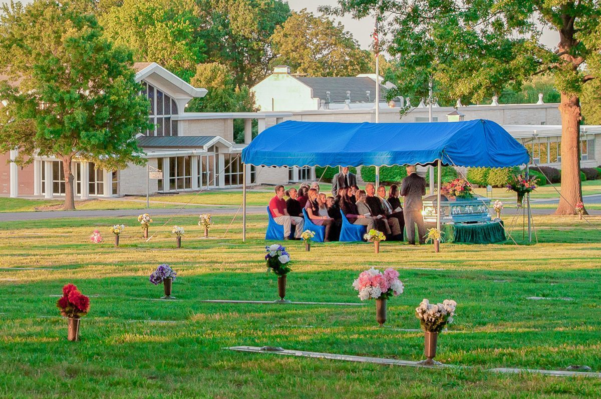 People seated under a blue canopy in a grassy park, with colorful flowers in the foreground.