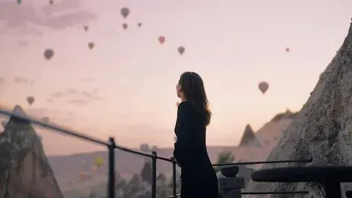 Silhouette of a person overlooking a pastel sunset sky with hot air balloons near rocky cliffs
