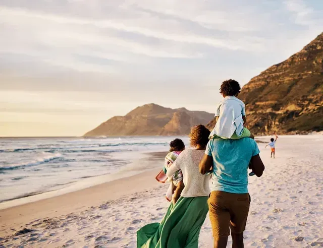 People walking along a sandy beach at sunset, with mountains and ocean waves in the background