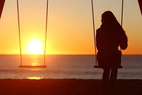 Silhouette on a swing at sunset over the ocean, with warm orange sky and sun near the horizon