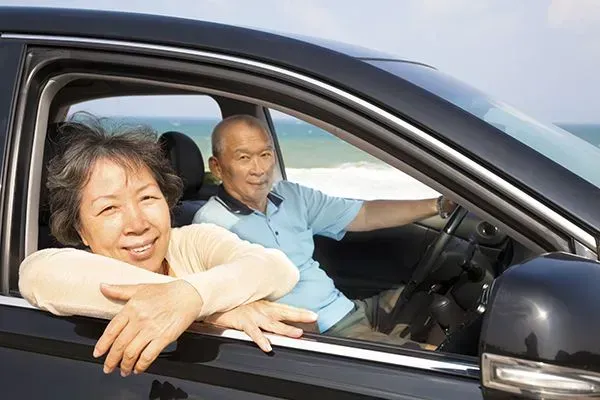 Two people smiling in a car, with one leaning out the window by the beach.