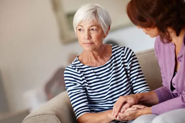 Two women sit on a couch, one holding the other’s hands in a supportive conversation.