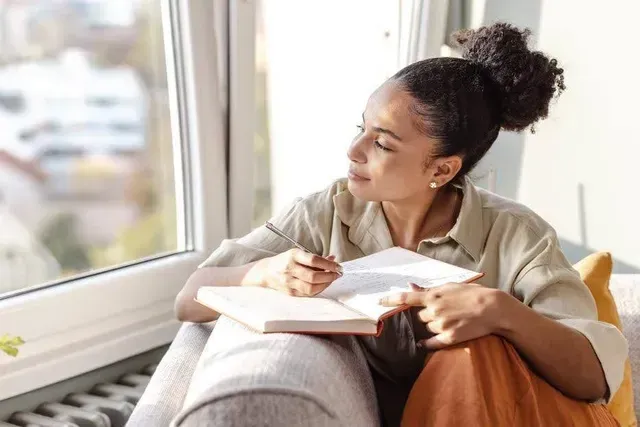 Person sitting by a window, writing in a notebook on a couch, looking thoughtful.