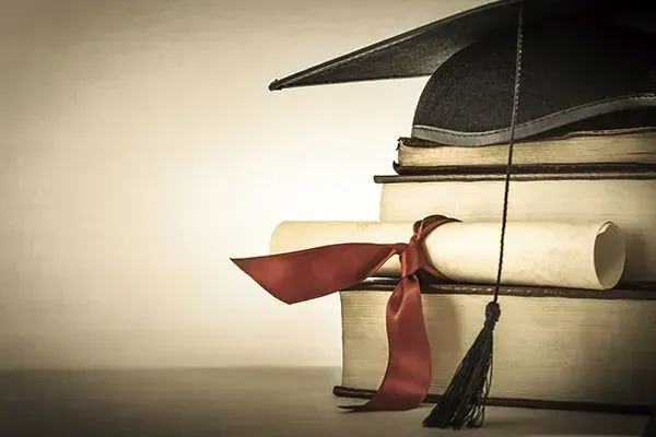 Graduation cap, diploma scroll, and red ribbon on stacked books against a warm background
