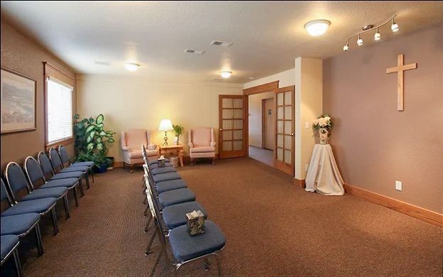Small chapel or prayer room with rows of blue chairs, a cross on the wall, and soft lighting.