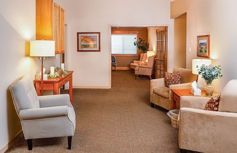 Quiet waiting room with beige chairs, a lamp, wall art, and a window into an adjoining room.