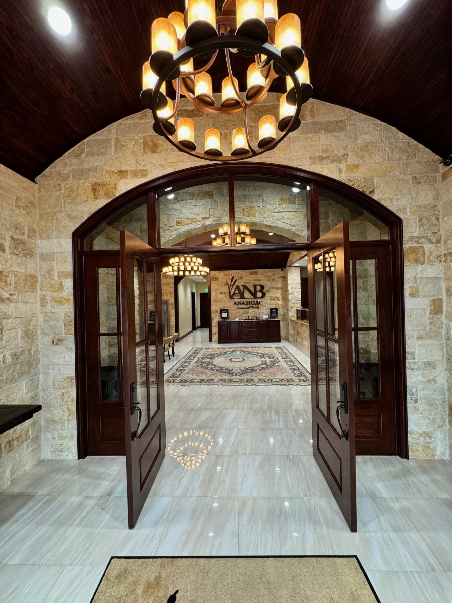 Open wooden doors in a stone hallway with a chandelier, leading to a patterned interior room