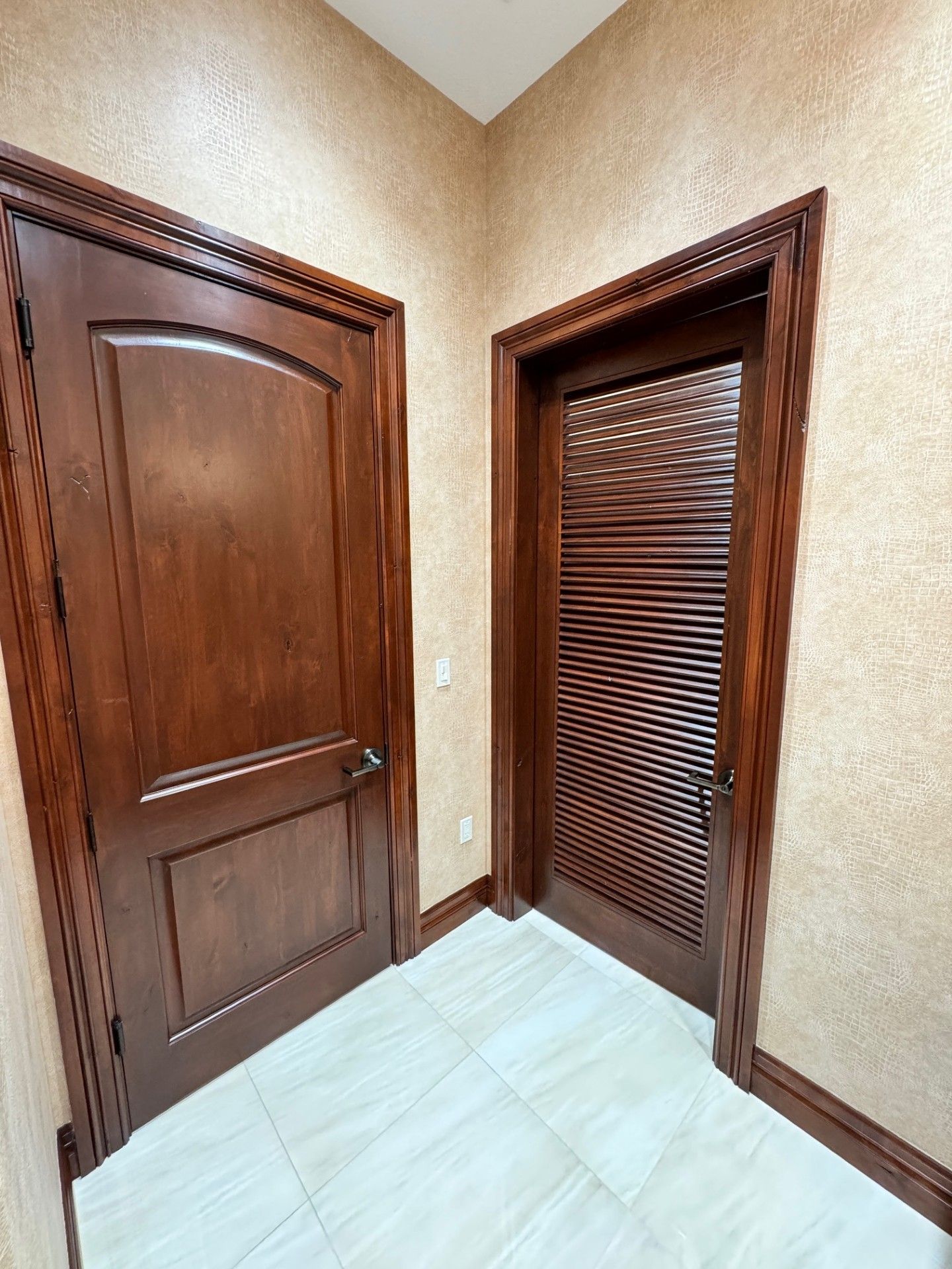 Small corner hallway with two wooden doors and beige textured walls, lit by natural light.