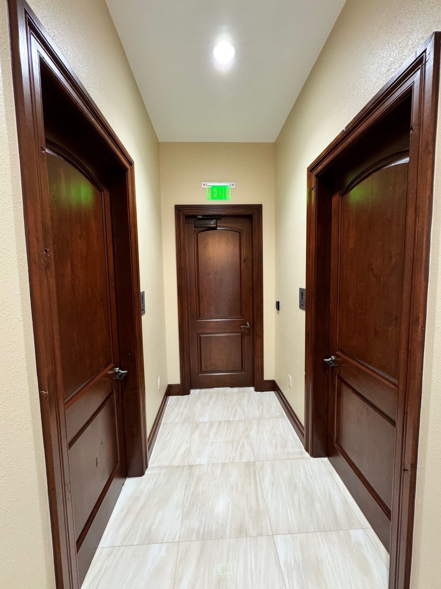 Narrow hallway with dark wooden doors, beige tile floor, and a lit exit sign above the far door