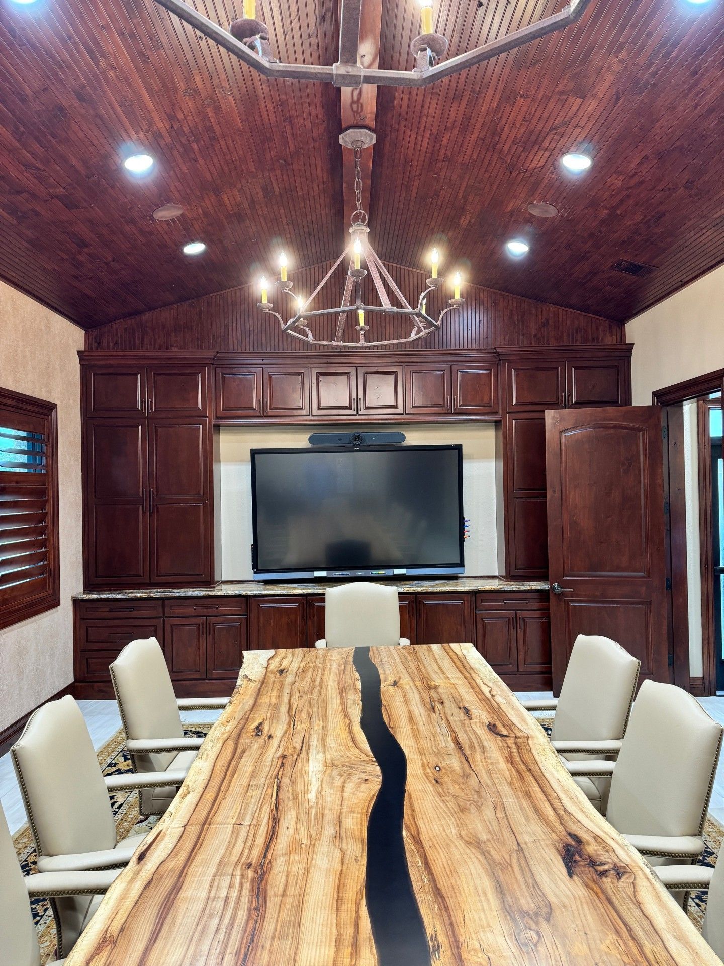 Elegant wood-paneled conference room with a live-edge table, white chairs, chandelier, and large TV screen