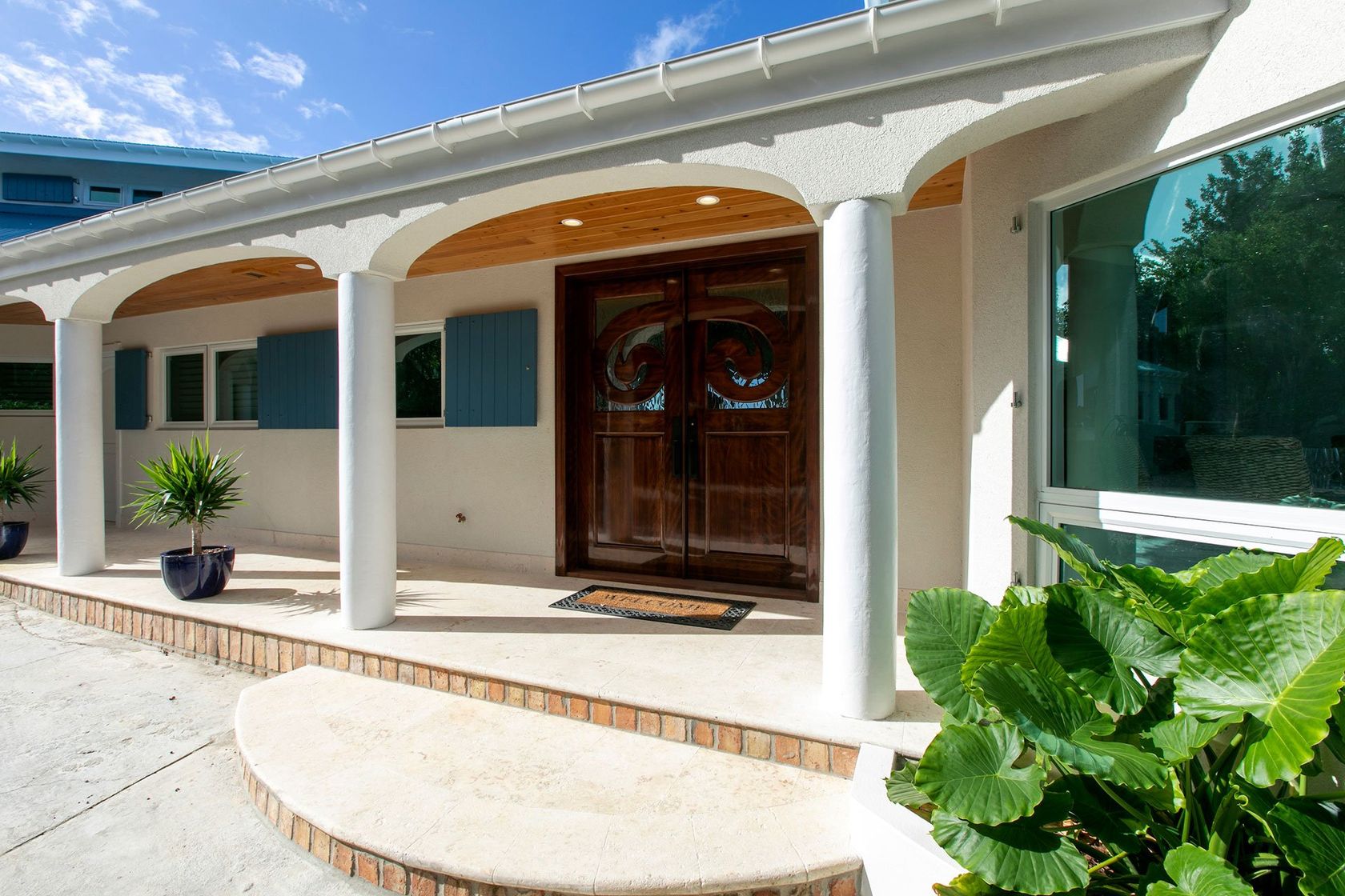 White house porch with columns, wooden double doors, and potted plants beside a bright window.