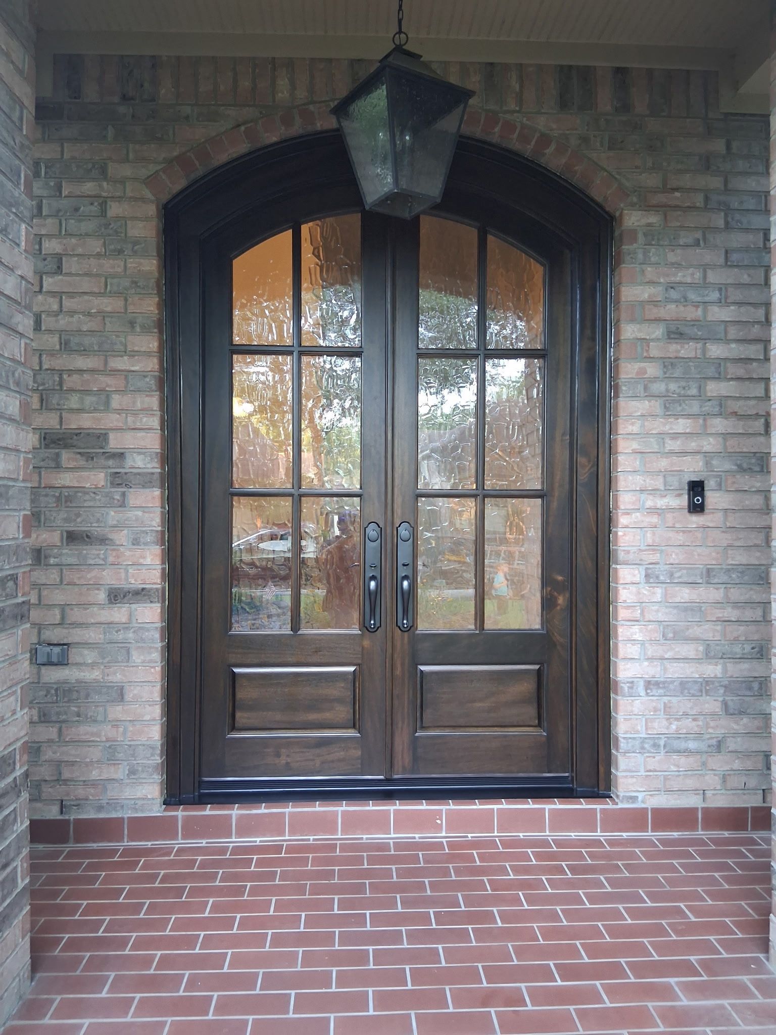 Brick-front entry with arched double wooden doors and a hanging lantern over a red brick porch
