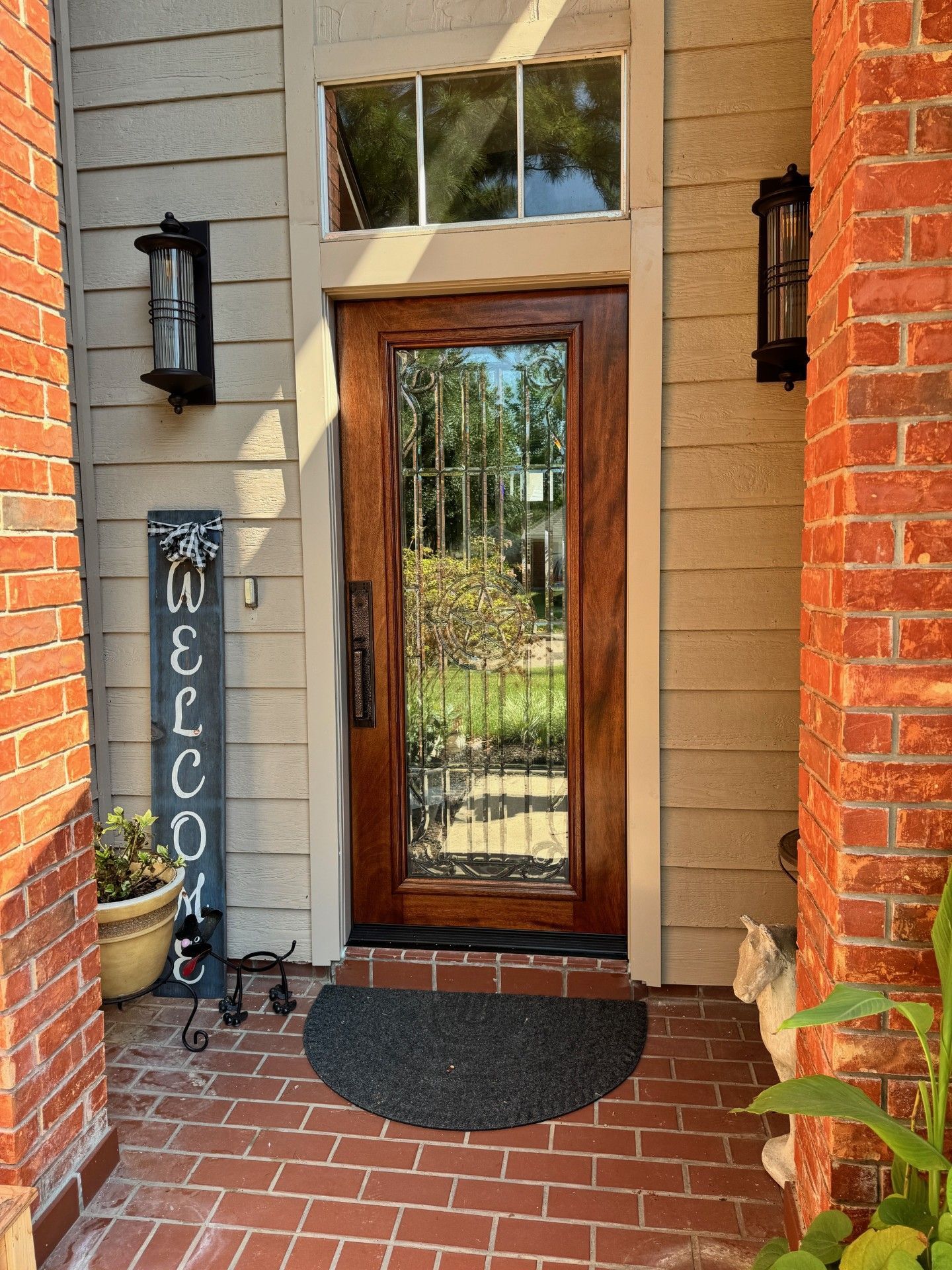 Front porch with a glass-paneled front door, brick walkway, wall lanterns, and a welcome sign.