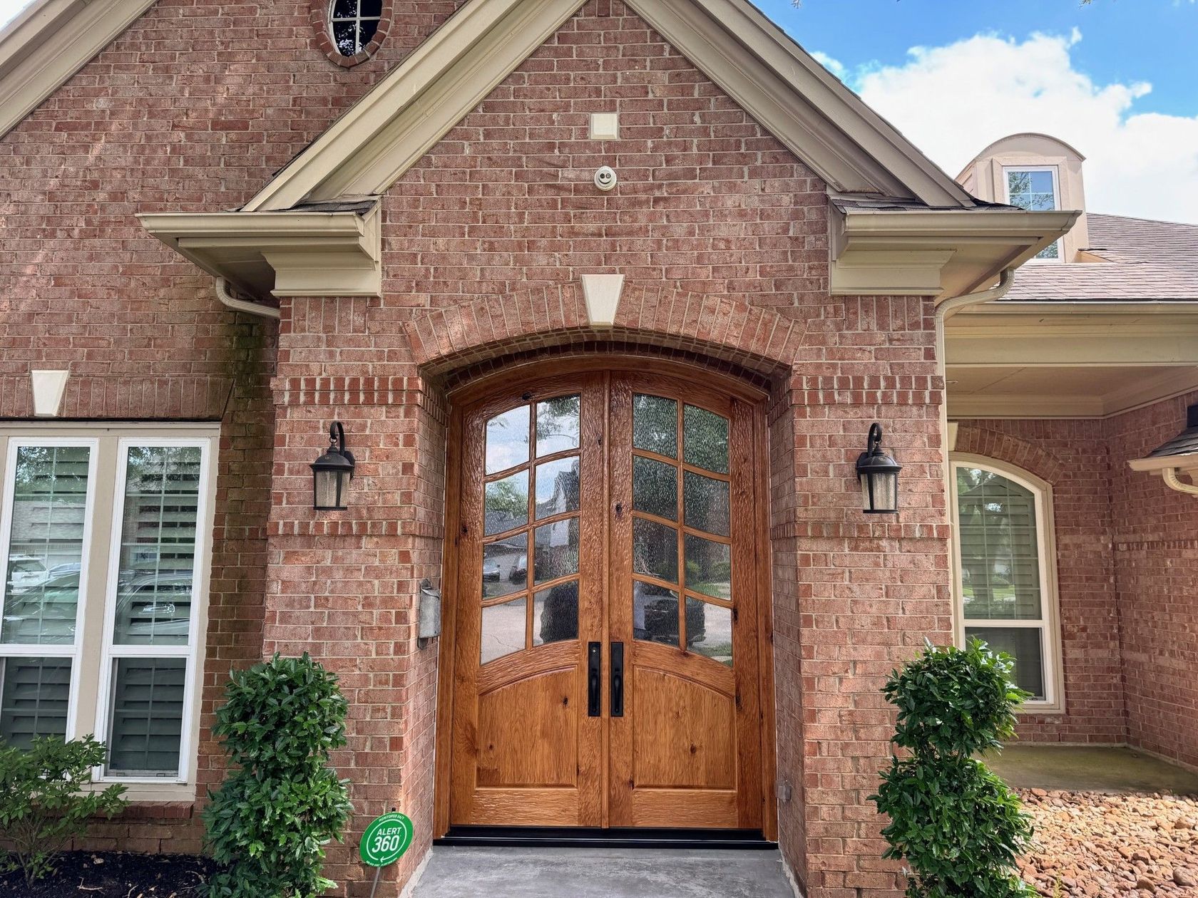 Brick house front entrance with double wooden arched doors, lanterns, and trimmed shrubs