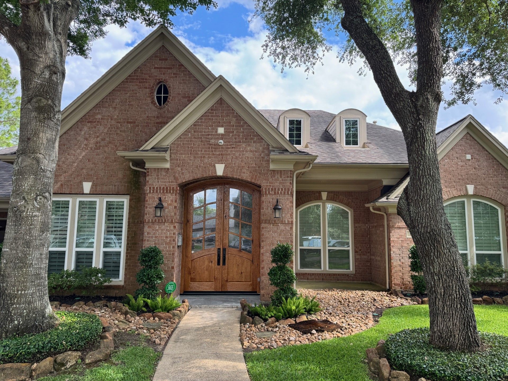 Brick house with arched wooden front door, white-trimmed windows, and a curved walkway under large trees