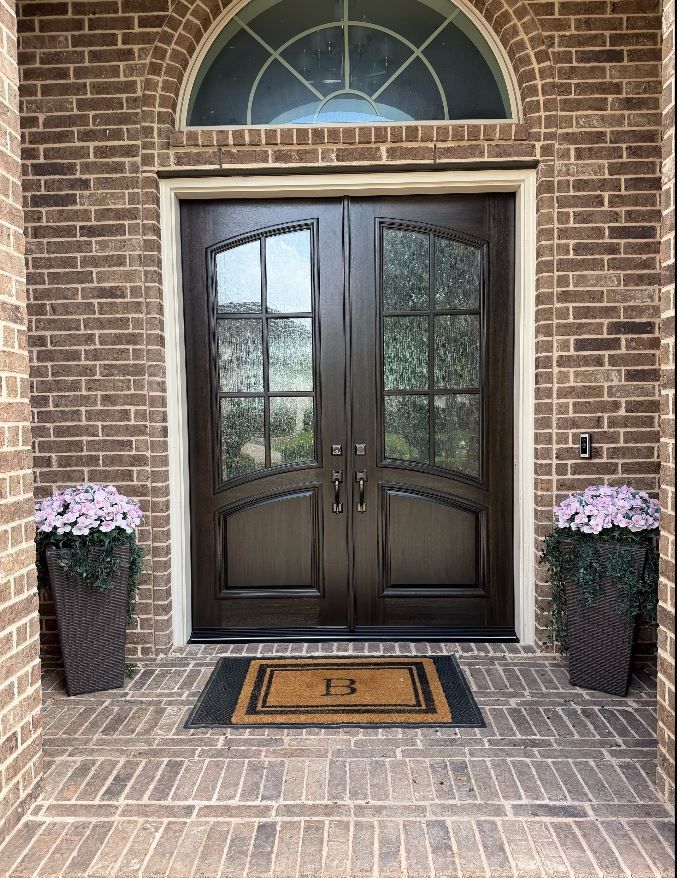 Brick front entry with double wooden doors, arched window, and two flower pots on the porch.
