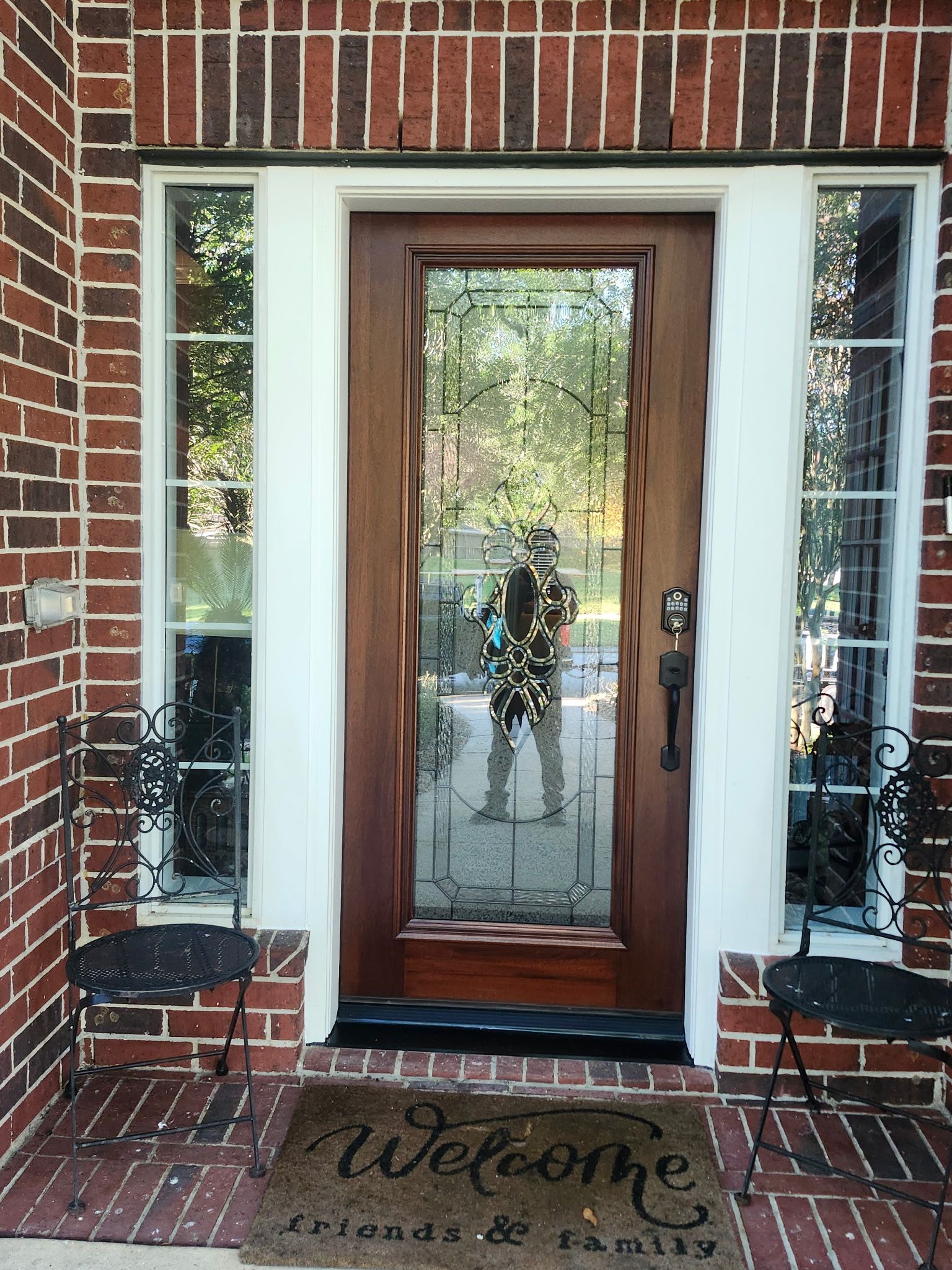 Front door with glass panels and brick entryway, flanked by narrow windows and a “Welcome” mat.