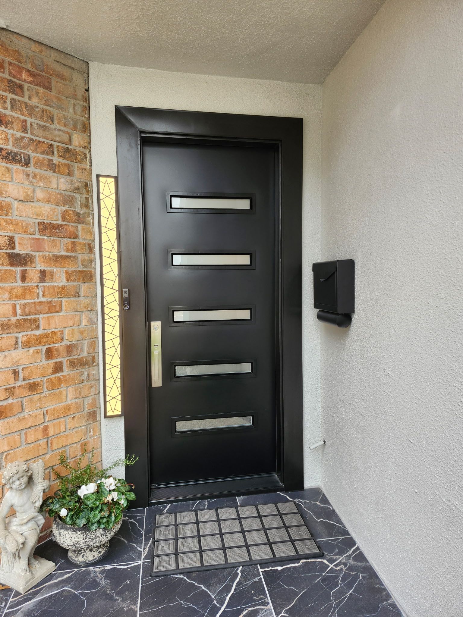 Black front door with narrow horizontal windows, a doormat, potted flowers, and a wall-mounted mailbox by a brick entryway
