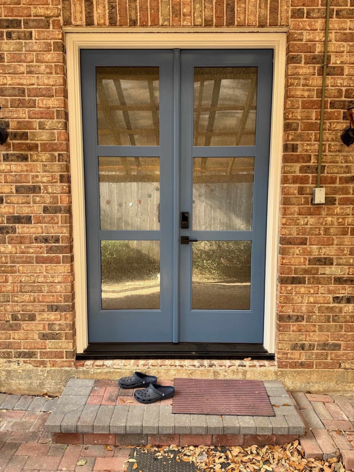 Blue double doors on a brick wall with a doormat and shoes on the doorstep.