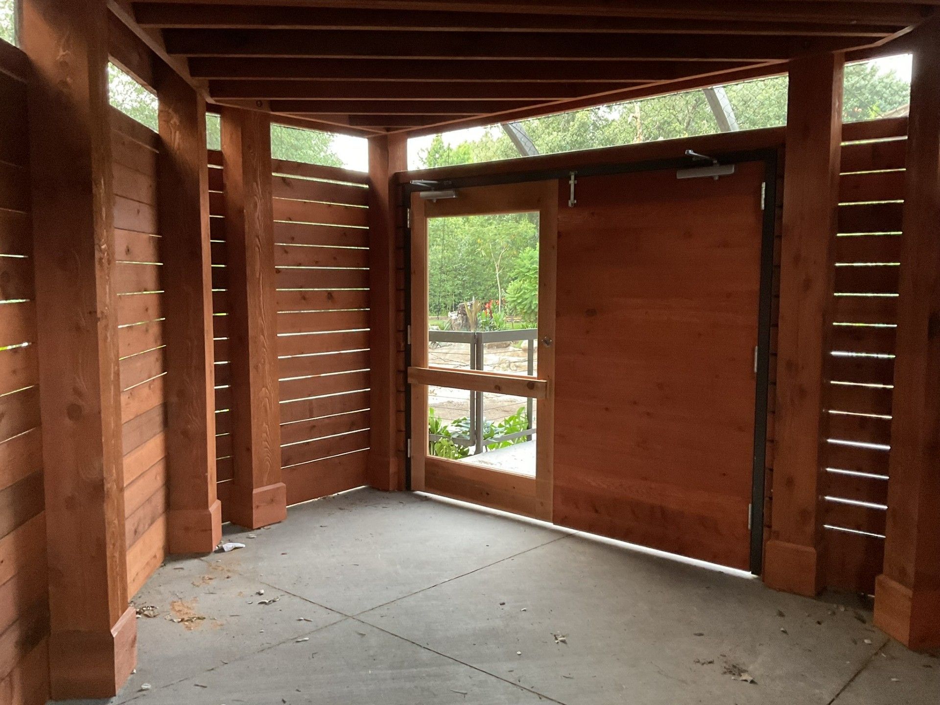 Empty wooden cabin interior with slatted walls, concrete floor, and a small window opening to greenery