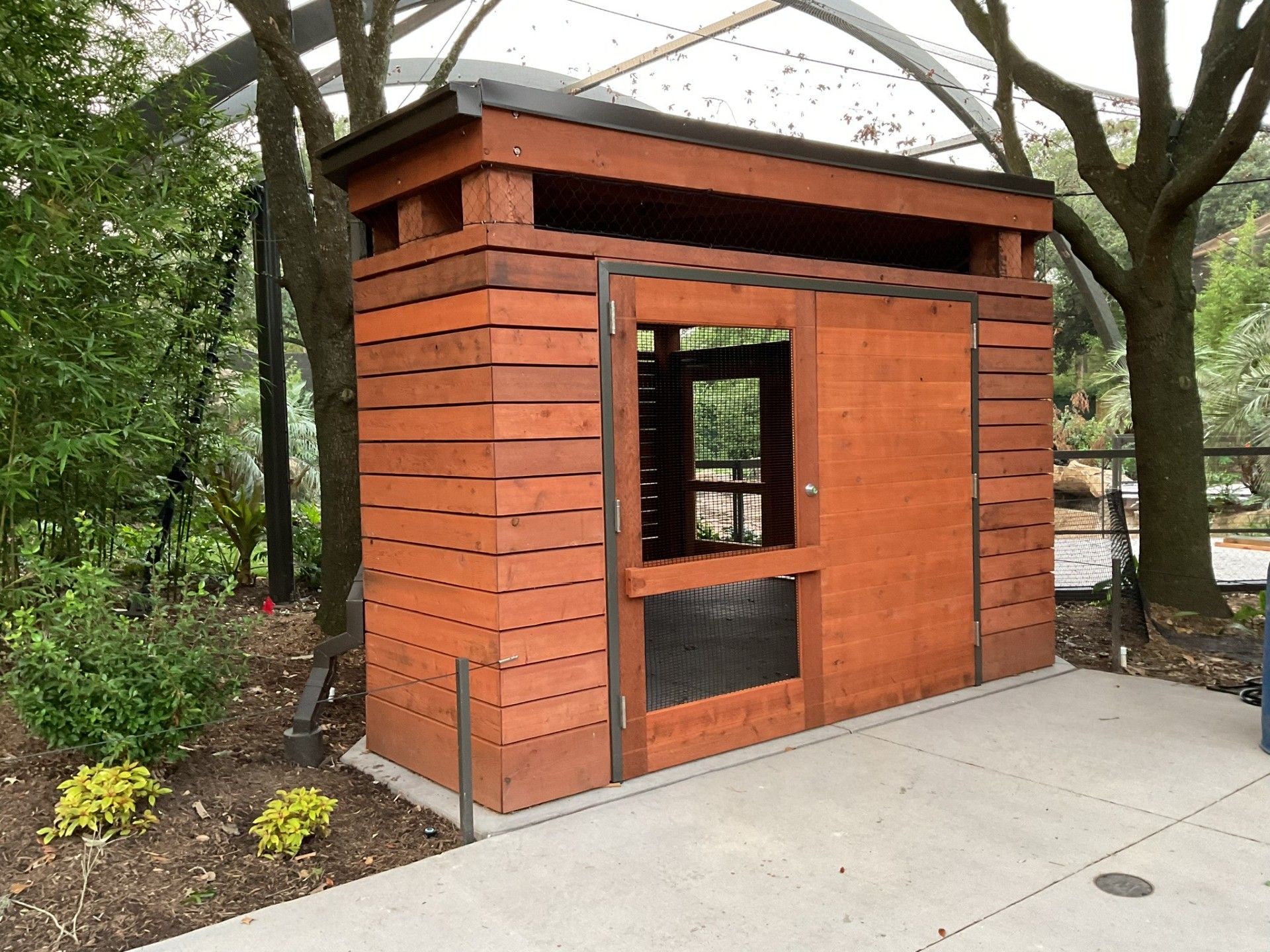 Small orange wooden kiosk or shed with a window, set beside a concrete path under trees.