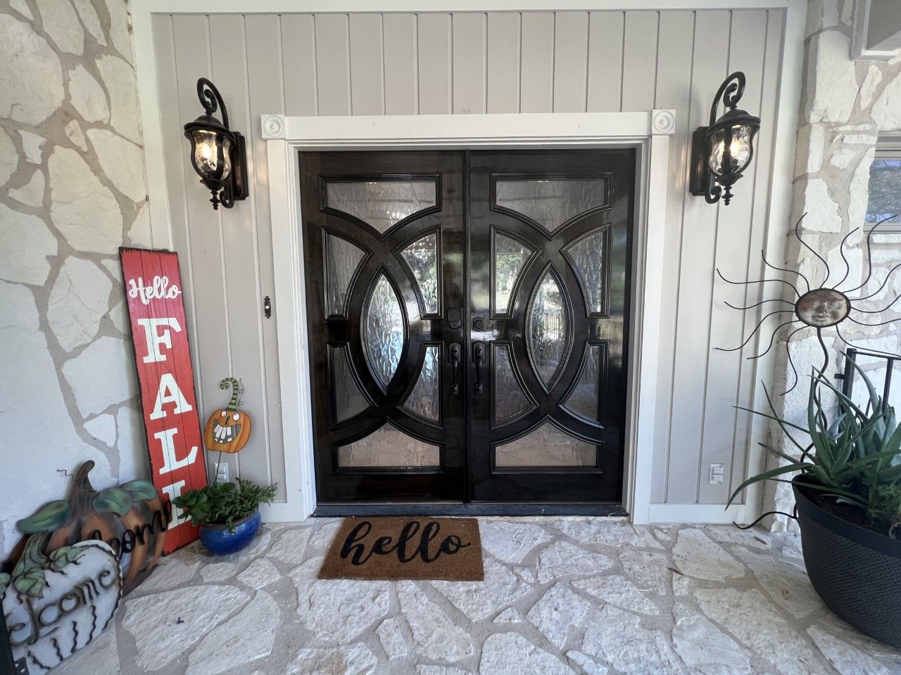 Front porch with black double doors, wall lanterns, a welcome mat, and fall decor including pumpkins and a “fall” sign