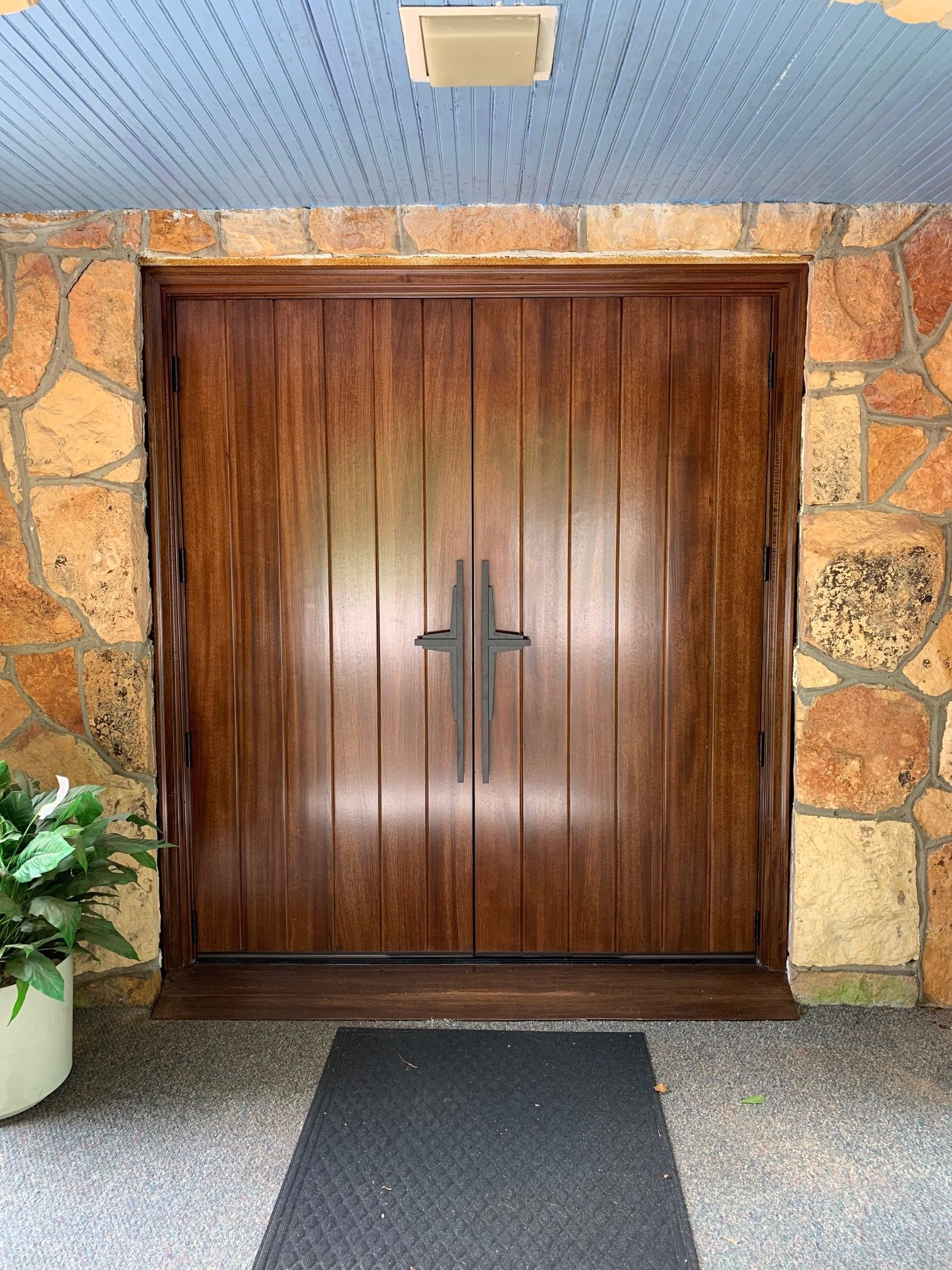 Wooden double doors in a stone entryway with a dark doormat and potted plant