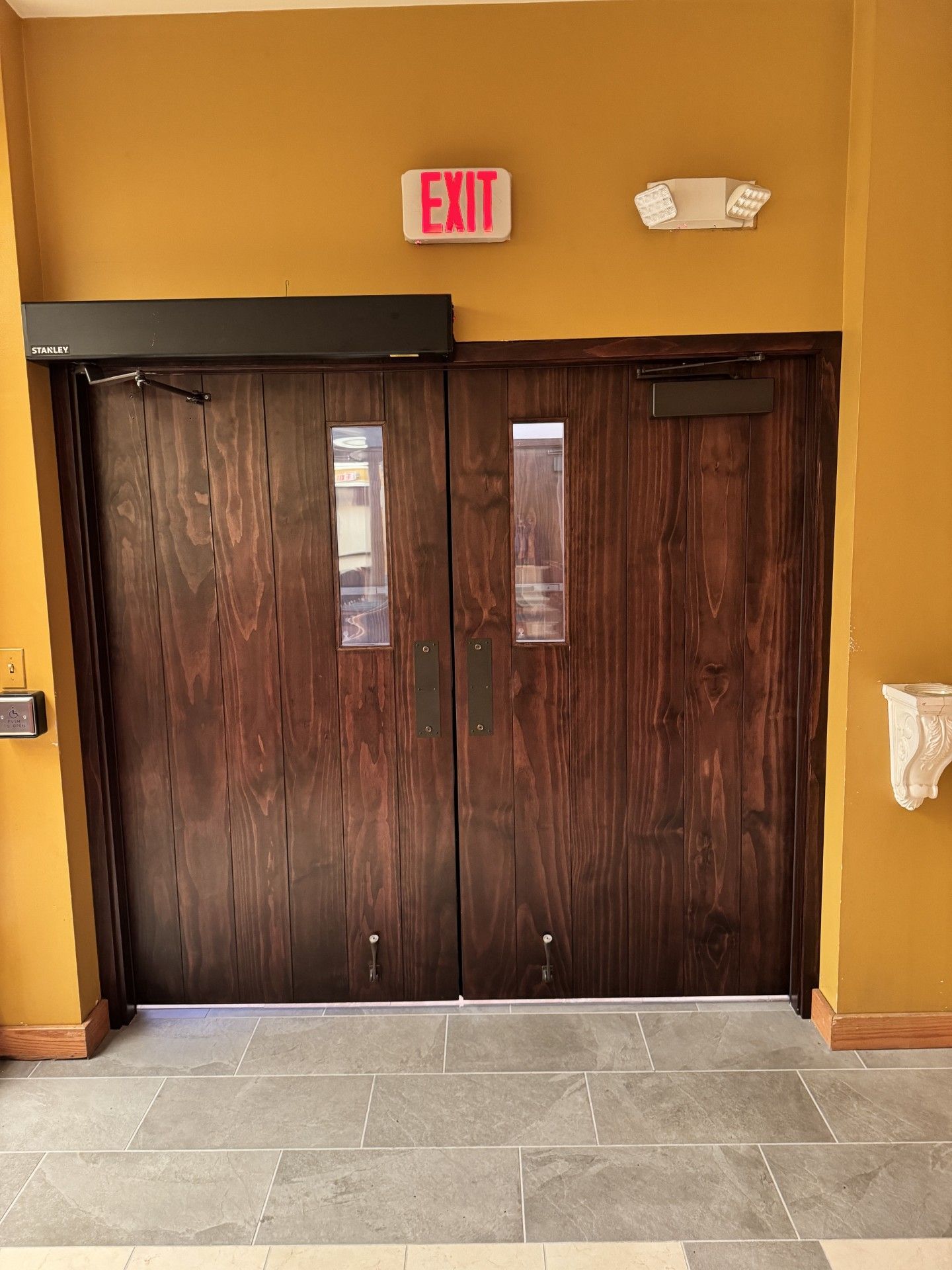 Closed dark wooden double doors beneath an EXIT sign in a tan hallway with tiled floor