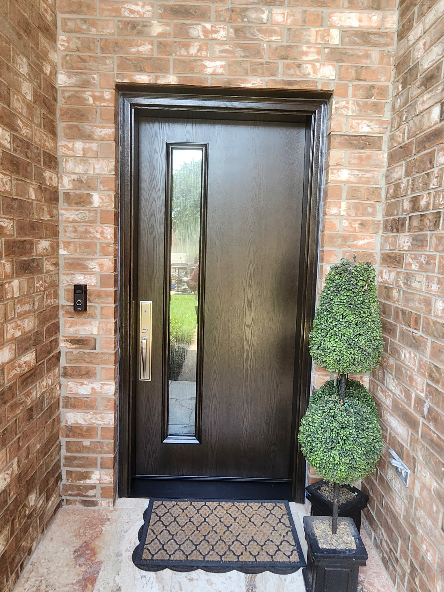 Black front door with narrow glass panel, set in a brick entryway beside a potted topiary.