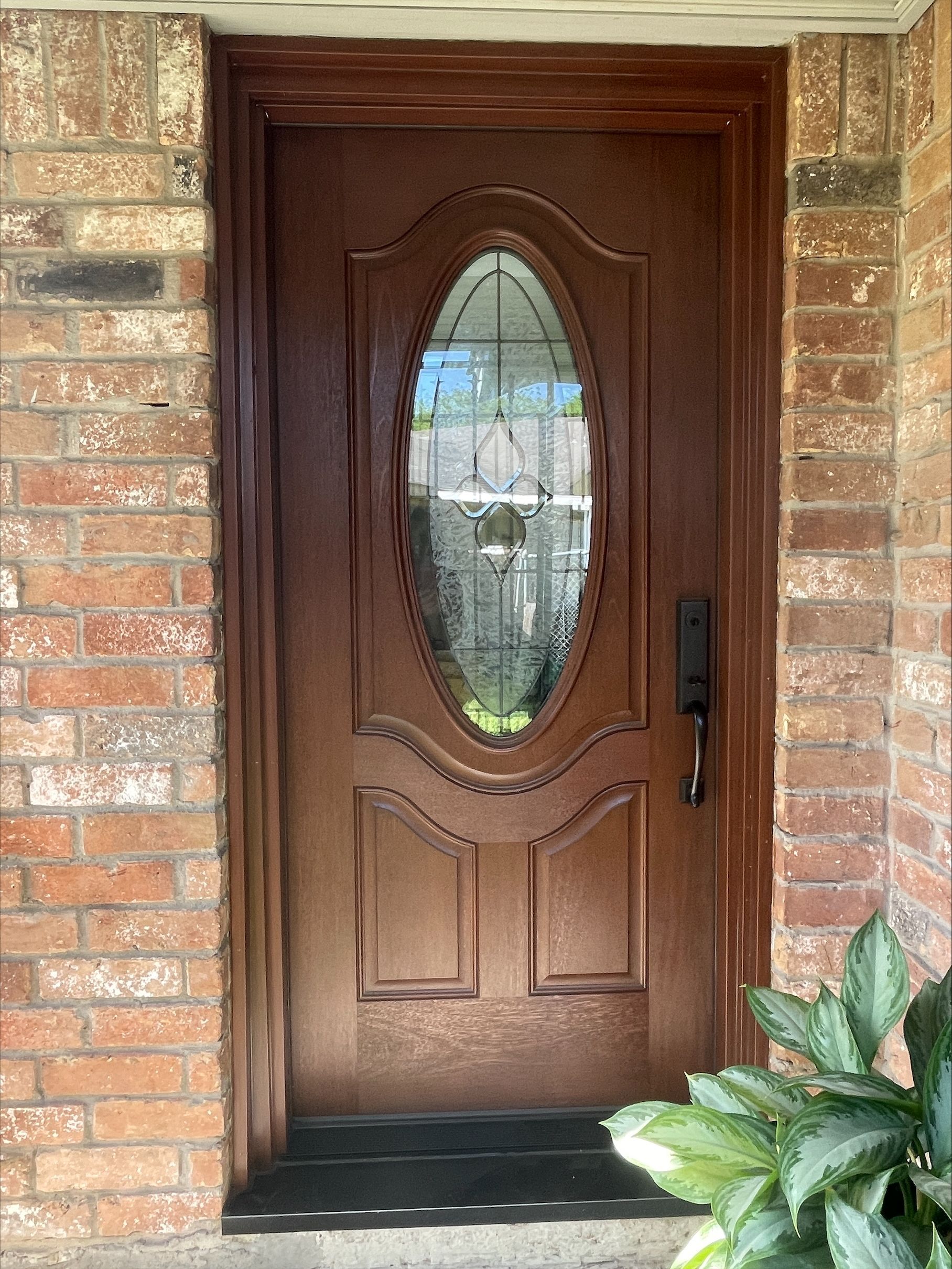 Brown front door with oval glass inset, brick exterior, and a potted plant beside the entrance