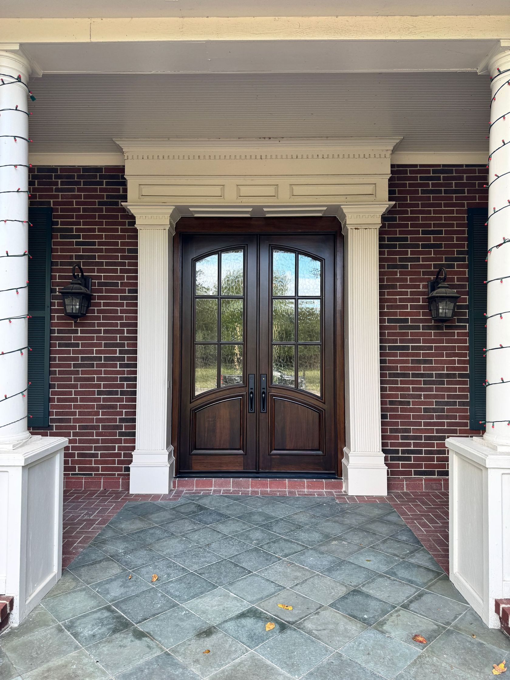 Front entrance with double wooden doors, white columns, and brick walls under a covered porch.