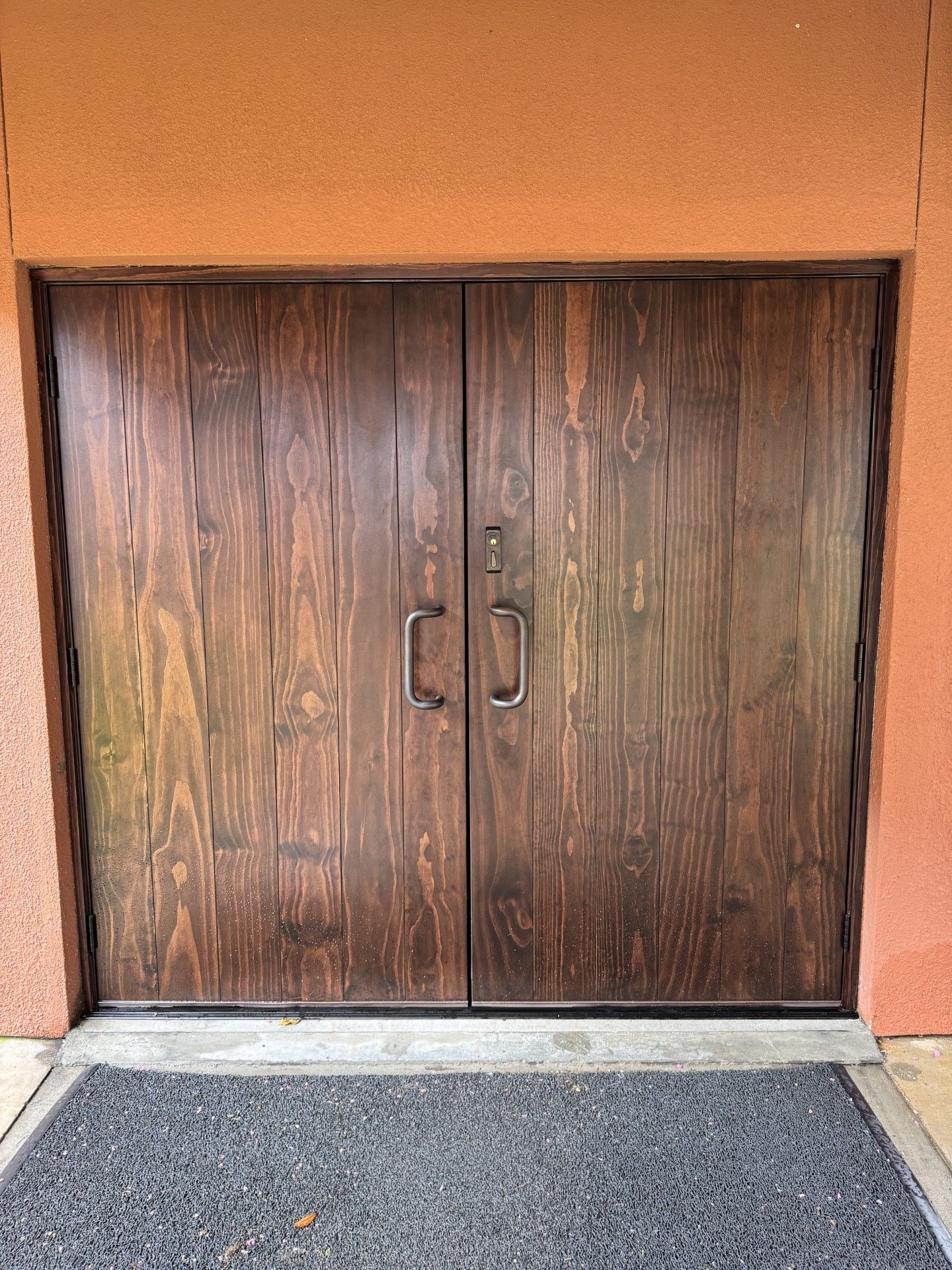 Wooden double doors set in an orange stucco wall above a dark entry mat.